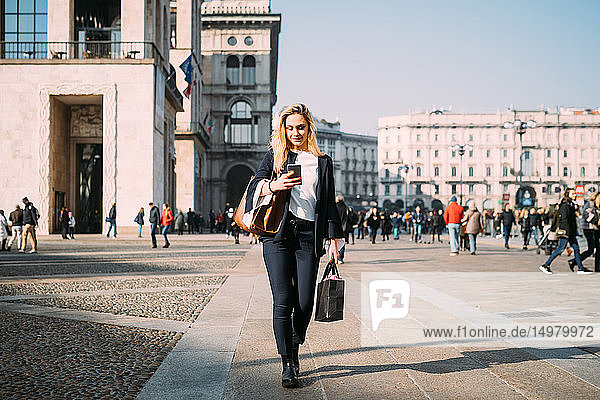 Young female tourist with shopping bags strolling and looking at smartphone in city square  Milan  Italy
