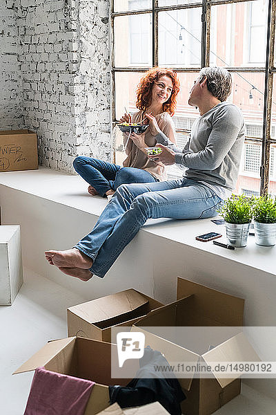 Ehepaar zieht in eine Wohnung im Industriestil sitzt am Fensterbrett und isst Essen zum Mitnehmen