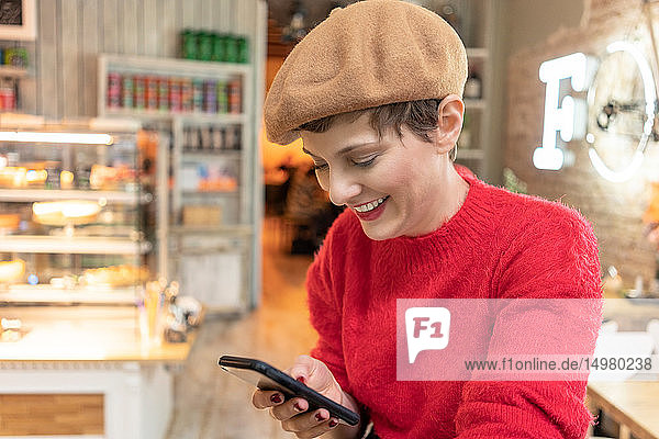 Mittelgroße erwachsene Frau mit Baskenmütze  die im Cafe ein Smartphone betrachtet