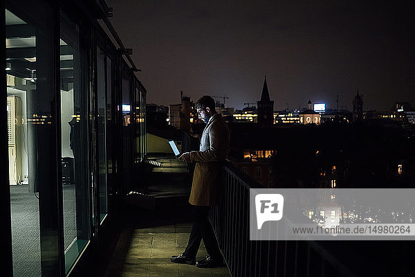 Mittlerer erwachsener Geschäftsmann schaut nachts auf den Laptop auf dem Balkon seines Büros