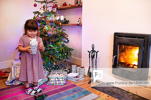 Mädchen steht neben dem Wohnzimmer-Weihnachtsbaum und betrachtet Weihnachtsgeschenk