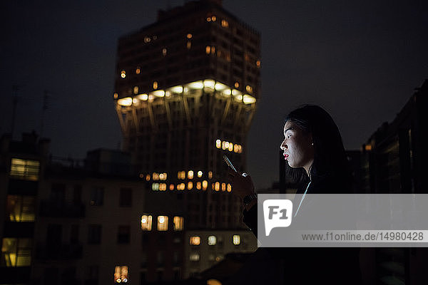 Junge Geschäftsfrau sieht nachts auf dem Balkon des Büros nach einem Smartphone