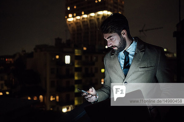 Mittlerer erwachsener Geschäftsmann schaut nachts auf Smartphone auf dem Balkon des Büros