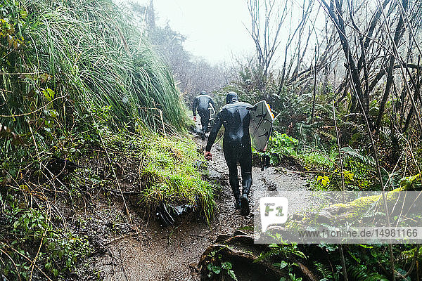 Young male surfers in wetsuits walking up coastal dirt track in rain  Arcata  California  United States