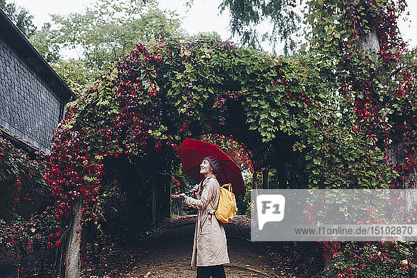 Frau hält Regenschirm an einem mit Reben bedeckten Bogen