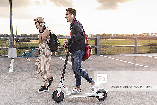 Happy young couple with cell phone  longboard and electric scooter on parking deck