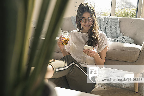 Junge Frau sitzt zu Hause mit Getränk und Handy auf dem Boden