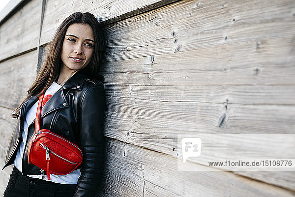 Glückliche junge Frau mit einer roten Hüfttasche  die an einer Holzwand lehnt