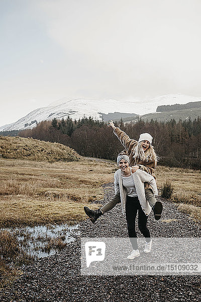 UK  Scotland  happy young woman carrying friend piggyback in rural landscape