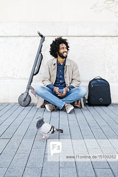 Lächelnder Mann sitzt auf seinem E-Scooter auf dem Bürgersteig  während im Vordergrund eine Taube vorbeizieht