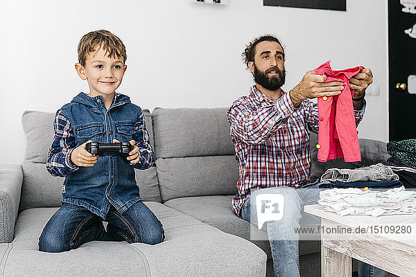 Porträt eines lächelnden kleinen Jungen beim Computerspiel  während sein Vater Wäsche faltet