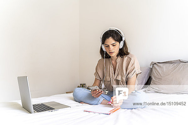 Junge Frau sitzt mit Kopfhörern und Laptop auf dem Bett und macht Notizen