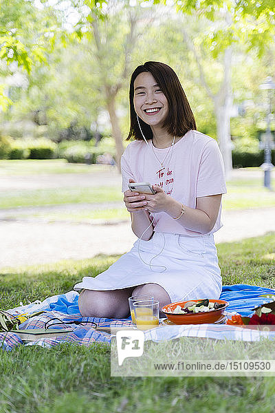 Glückliche junge Frau mit Handy und Kopfhörern beim Picknick im Park