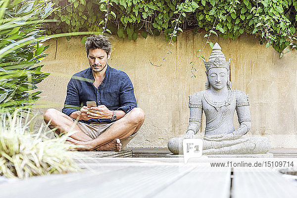 Mann sitzt im Schneidersitz neben der Buddhastatue in einem Zen-Garten und benutzt ein Smartphone
