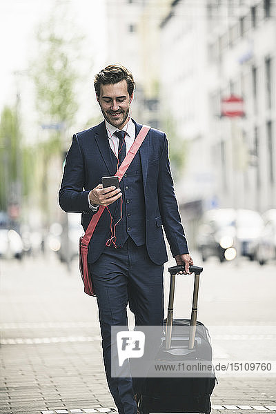 Smiling businessman walking in the city with cell phone and suitcase