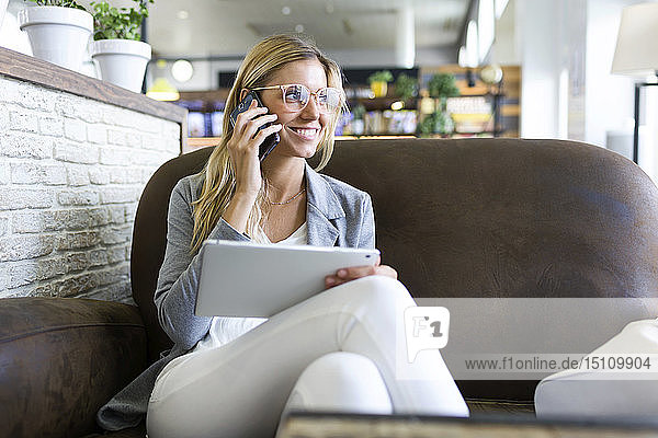 Junge Frau telefoniert mit ihrem Handy  während sie ihr digitales Tablet in einem Café benutzt