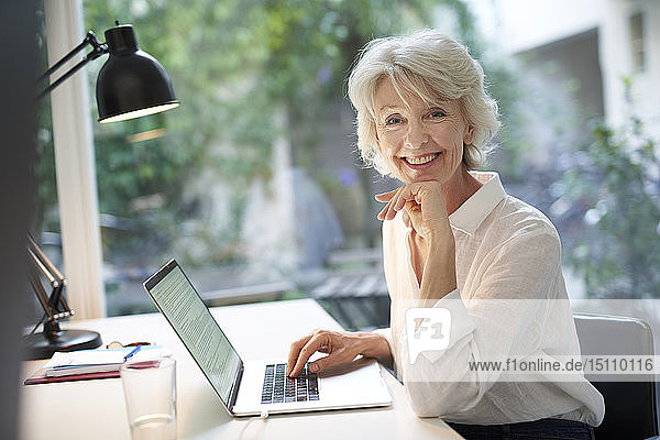Porträt einer lächelnden reifen Geschäftsfrau  die am Schreibtisch sitzt und einen Laptop benutzt