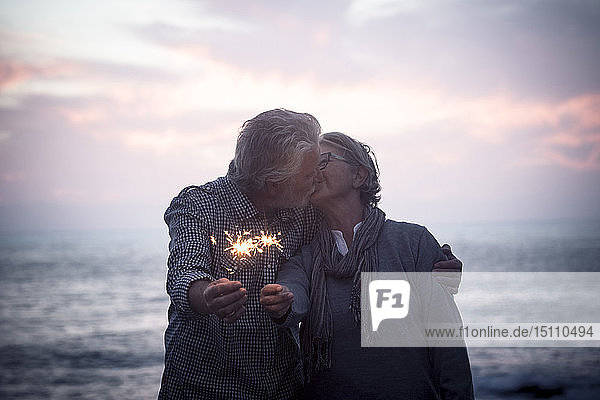 Küssendes älteres Paar  das bei Sonnenuntergang mit Wunderkerzen vor dem Meer steht