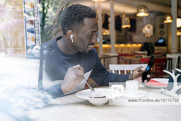Lächelnder junger Mann mit Kopfhörern und Smartphone hinter einer Fensterscheibe in einem Restaurant