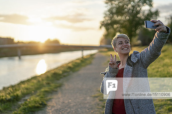 Porträt einer grinsenden Frau  die sich bei Sonnenuntergang mit einem Smartphone selbstständig macht