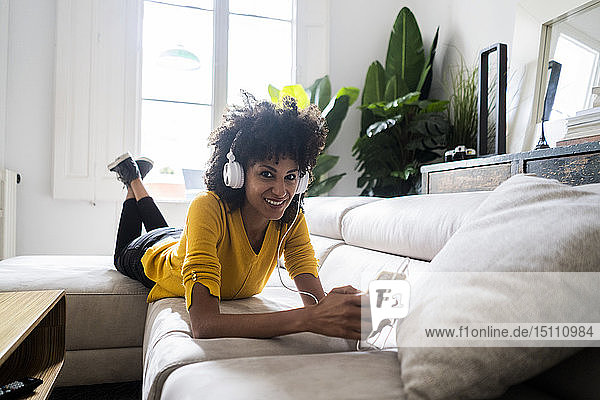 Porträt einer lächelnden Frau  die mit Handy und Kopfhörer auf der Couch liegt
