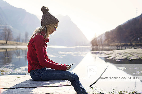 Junge blonde Frau sitzt im Winter auf einem Steg an einem See