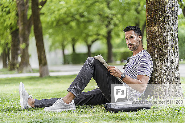 Man leaning against a tree in park reading newspaper
