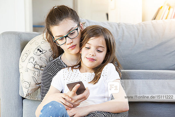 Zwei Schwestern sitzen vor der Couch und schauen auf das Smartphone