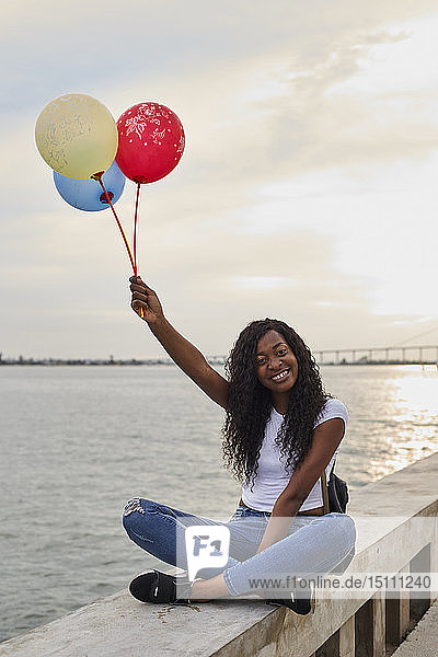 Mocambique  Maputo  Porträt einer glücklichen jungen Frau mit drei Luftballons  die vor dem Meer sitzt
