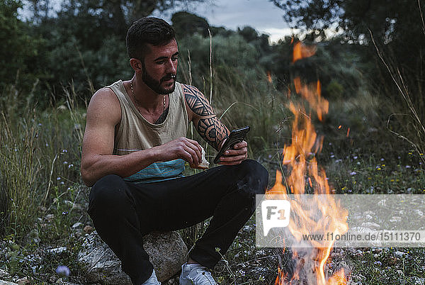 Mann sitzt mit Handy am Lagerfeuer in ländlicher Landschaft