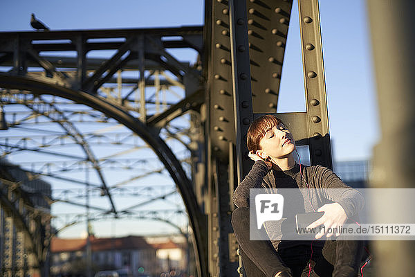 Porträt einer entspannten jungen Frau  die auf einem Stahlträger einer Brücke sitzt und mit einem Smartphone Musik hört