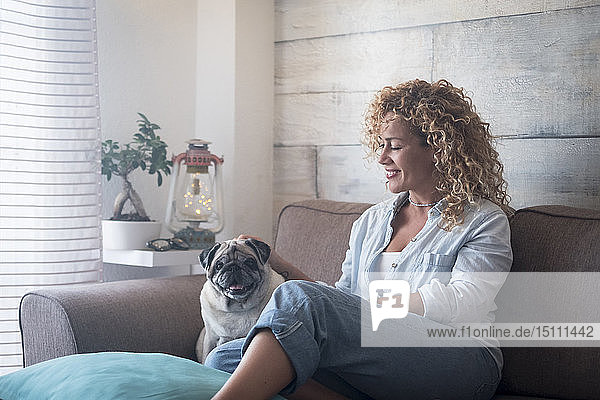 Woman sitting on couch at home petting pug dog