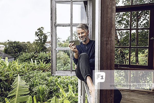 Mann mit Handy am Fenster sitzend in tropischer Umgebung