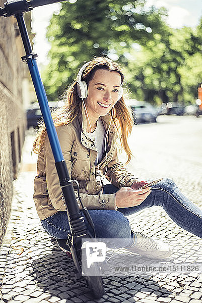 Porträt einer lächelnden Frau  die auf einem E-Scooter sitzt und mit Kopfhörern und Smartphone Musik hört