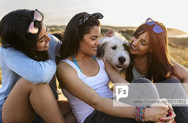 Drei glückliche Frauen mit Hund sitzen bei Sonnenuntergang in den Dünen