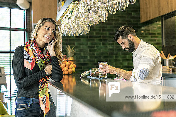 Lächelnde Frau am Handy an der Theke einer Bar mit Barkeeper  der einen Cocktail zubereitet
