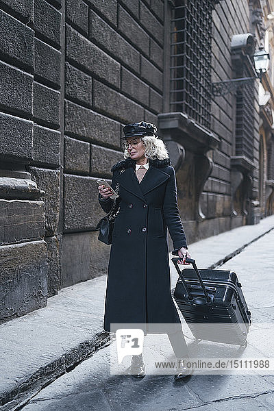 Reife Geschäftsfrau mit schwarzem Mantel und Ledermütze auf Geschäftsreise beim Blick auf Mobiltelefon