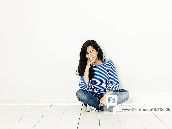Hübsche junge Frau mit schwarzen Haaren und blau-weiß gestreiftem Pullover  die vor weißem Hintergrund auf dem Boden sitzt