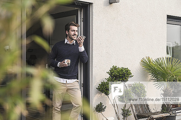 Junger Mann lehnt in Tür seines Hauses  hält Tasse Kaffee in der Hand  benutzt Smartphone