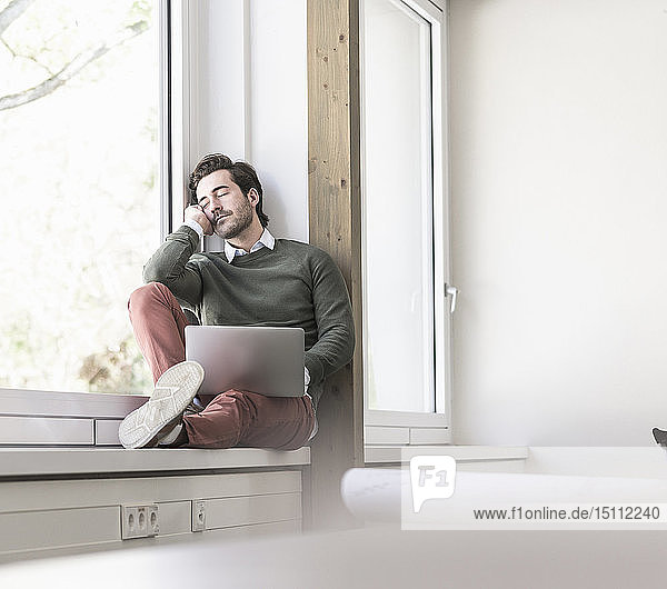 Junger Geschäftsmann mit Laptop sitzt auf der Fensterbank und macht eine Pause