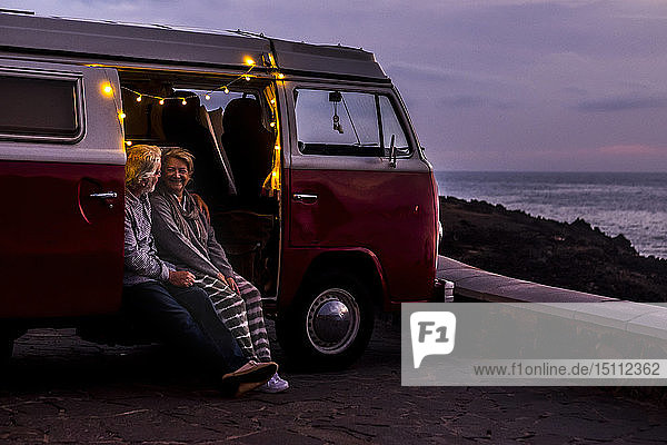 Älteres Ehepaar reist in einem Oldtimer-Van und betrachtet den Sonnenuntergang am Meer