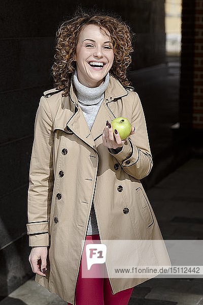 Bildnis einer lachenden Frau mit grünem Apfel in beigem Trenchcoat und Rollkragenpullover