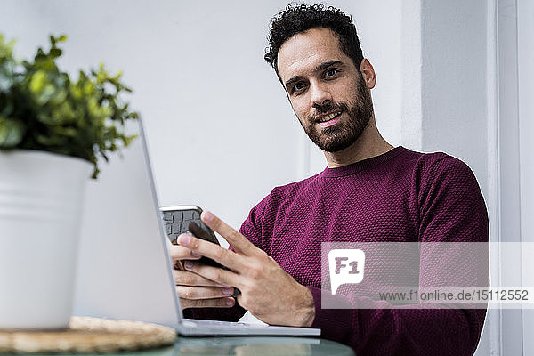 Porträt eines jungen Mannes  der zu Hause am Tisch sitzt und mit Laptop und Handy telefoniert