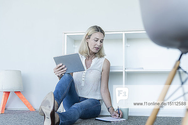 Lässige Geschäftsfrau  die im Büro auf dem Boden sitzt  ein Tablett benutzt und Notizen macht