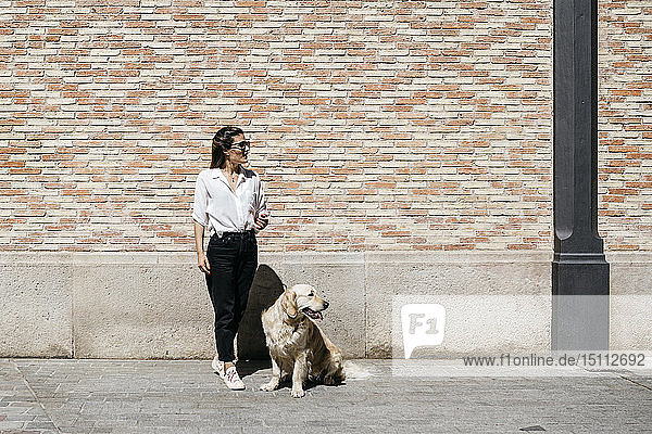 Frau und ihr Labrador-Retriever warten vor einer gemusterten Ziegelmauer und schauen in die Ferne
