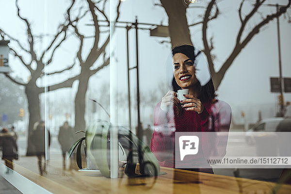 Lächelnde junge Frau mit einer Tasse Kaffee hinter einer Fensterscheibe in einem Cafe