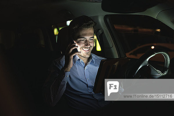 Lächelnder junger Mann benutzt nachts im Auto Handy und Tablet
