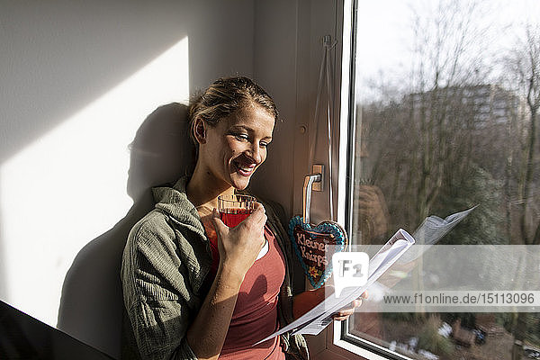 Lächelnde junge Frau liest Zeitung am Fenster