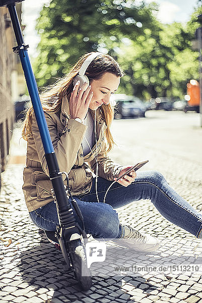 Lächelnde Frau sitzt auf einem E-Scooter und hört Musik mit Kopfhörern und Smartphone