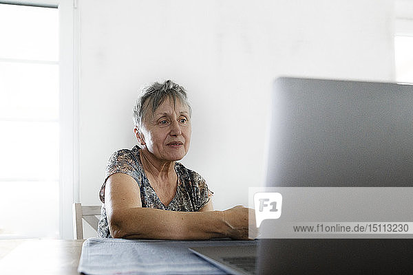Ältere Frau sitzt zu Hause am Tisch mit Laptop